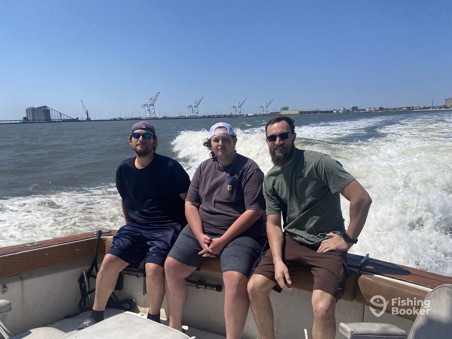 Three anglers enjoying a sunny day on a boat, with a view of the coastline and industrial backdrop.