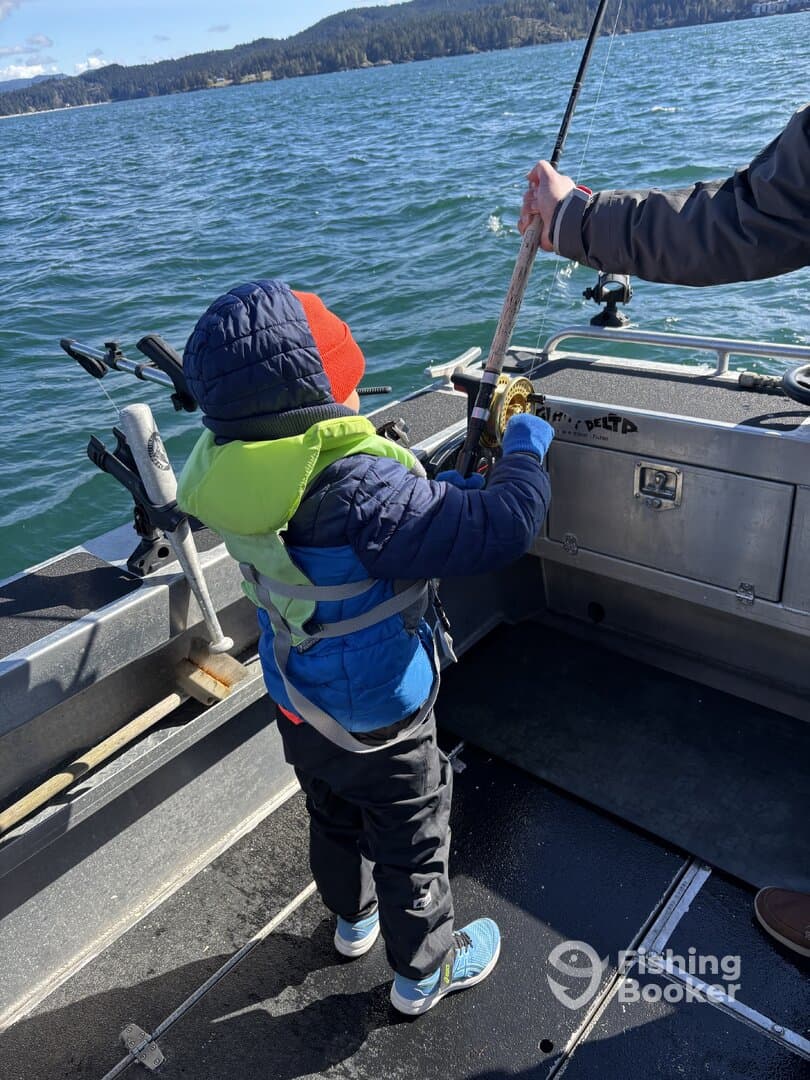 A young angler in a life jacket is actively reeling in a fishing line on a boat, surrounded by a scenic water backdrop.