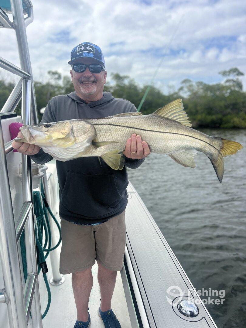 A proud angler displaying a large Snook caught while fishing on a boat, surrounded by a scenic waterway.