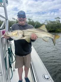 A proud angler displaying a large Snook caught while fishing on a boat, surrounded by a scenic waterway.