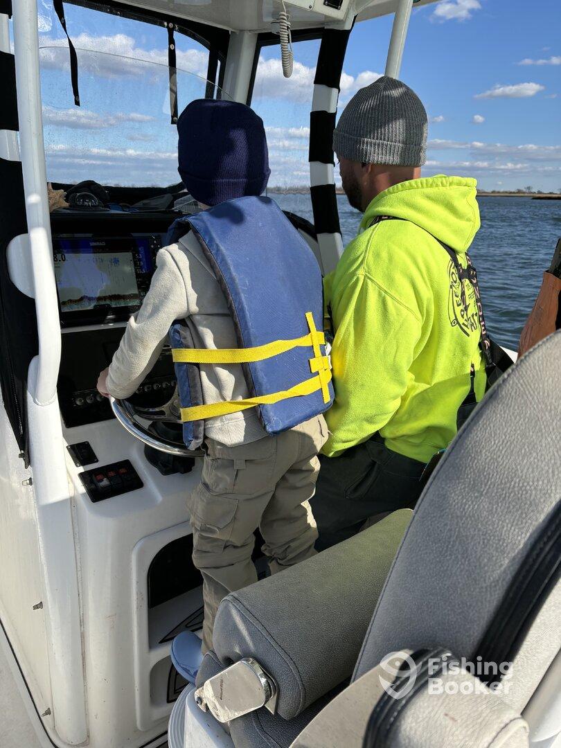 A young boy in a life jacket is steering the boat under the guidance of an adult, showcasing a family-friendly fishing experience.