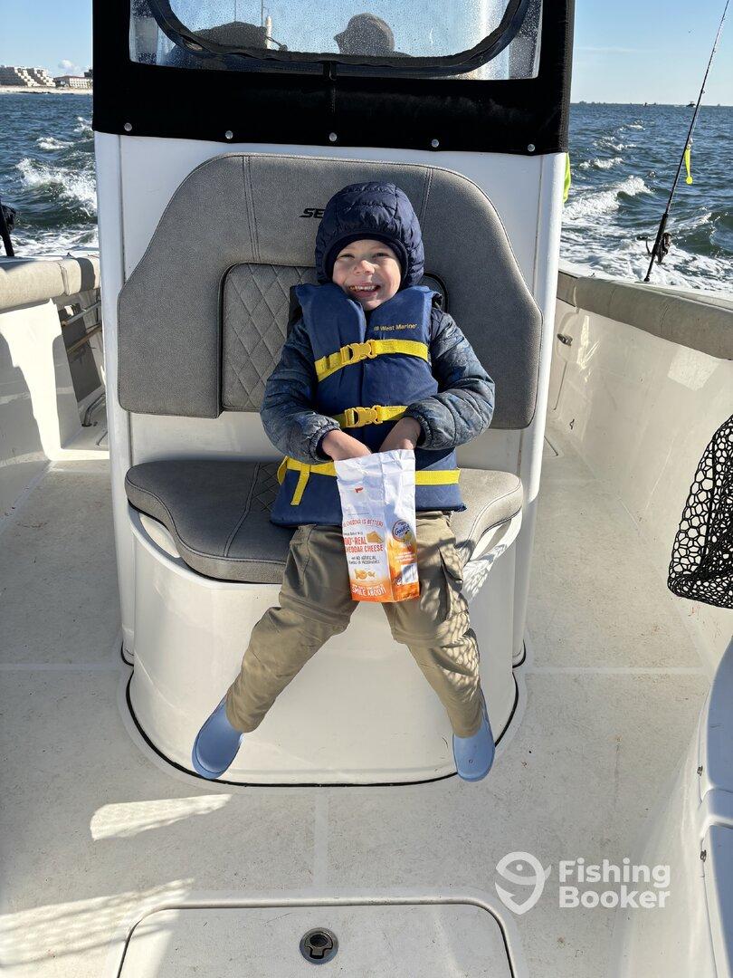 A young boy in a life jacket enjoying a snack while seated inside a fishing boat, showcasing a family-friendly fishing experience.