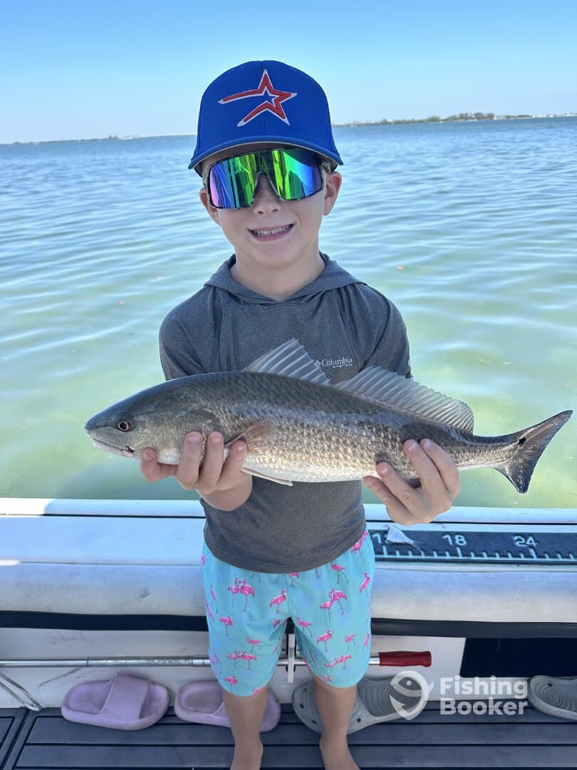 A young angler proudly displaying a Redfish while fishing on a boat in calm waters.