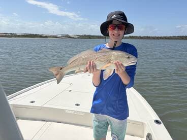 A young angler proudly displaying a Redfish caught while fishing on a boat in calm waters.