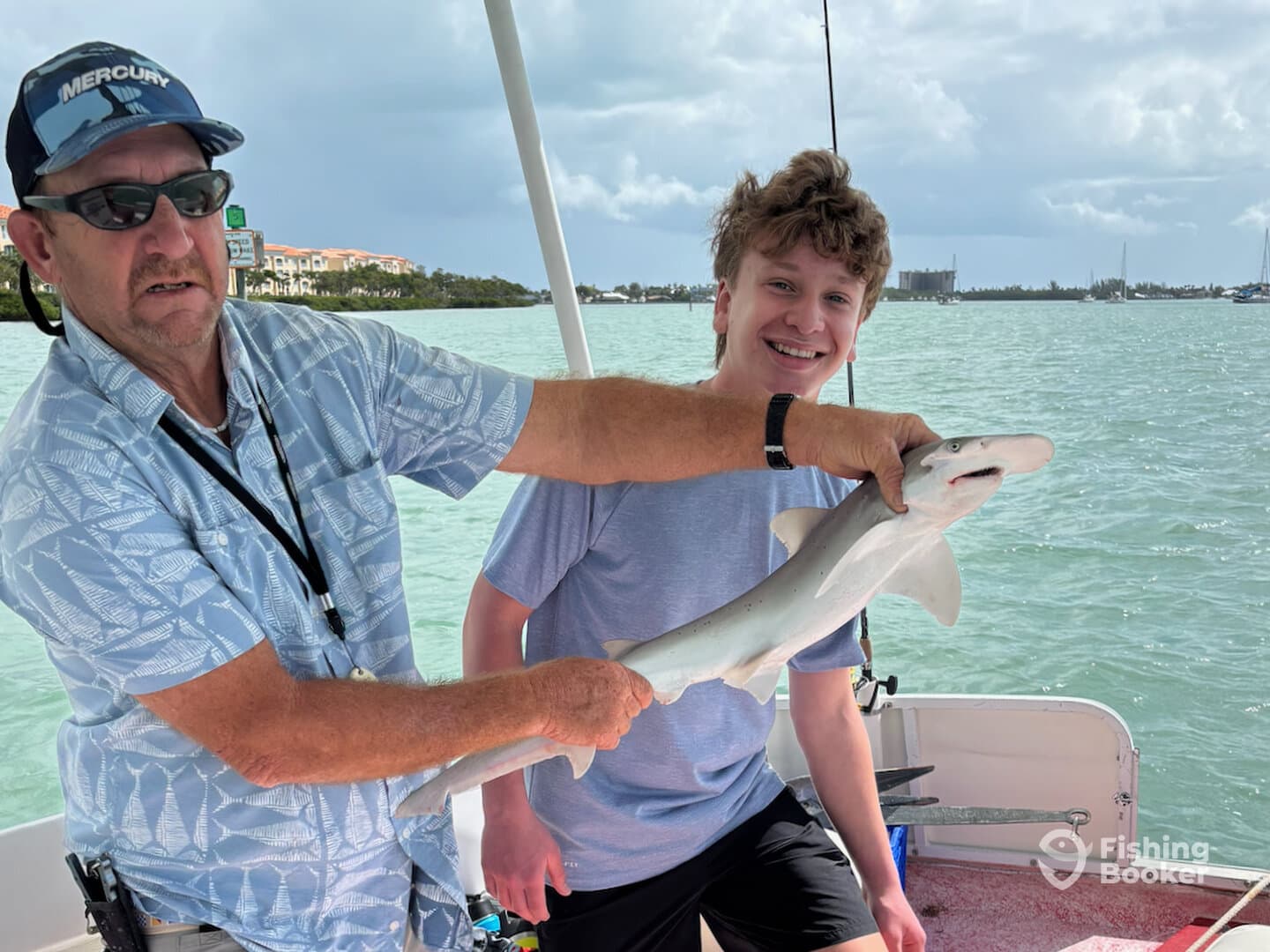 A young angler and an adult proudly displaying a caught shark while fishing on a boat in clear waters.