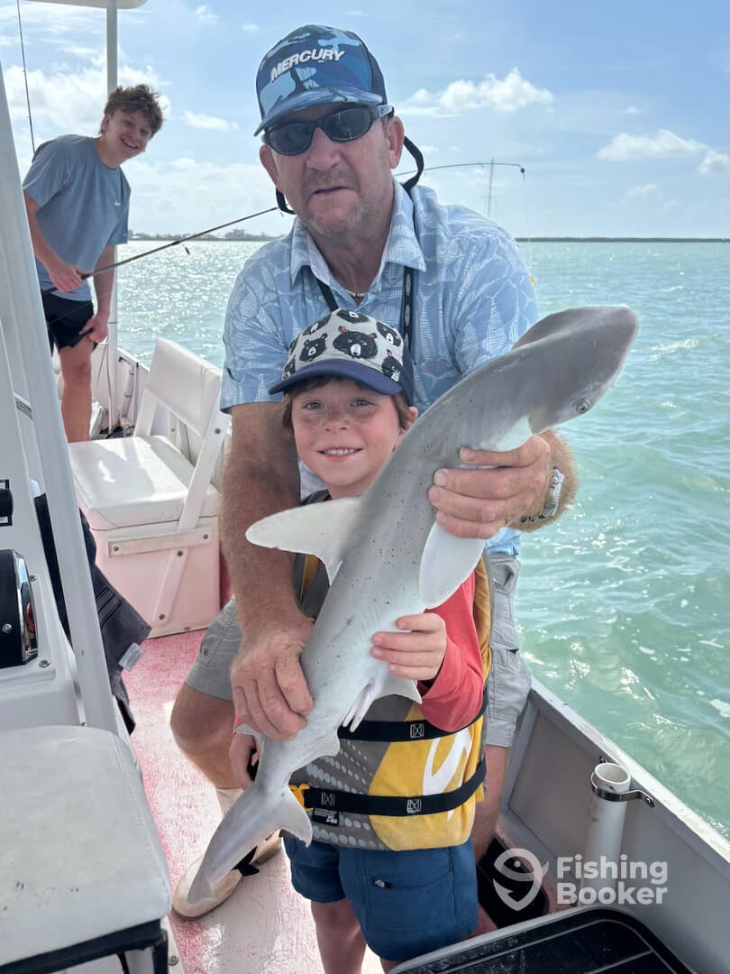 A young boy proudly displays a caught shark while fishing with family on a boat, enjoying a sunny day on the water.