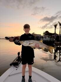 A young angler proudly displaying a large Snook while fishing at sunset from a boat.