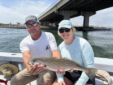 Two anglers proudly displaying a large Speckled Trout while fishing from a boat under a bridge.