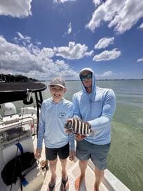A young angler and an adult proudly display a striped fish on a boat, enjoying a sunny day on the water.