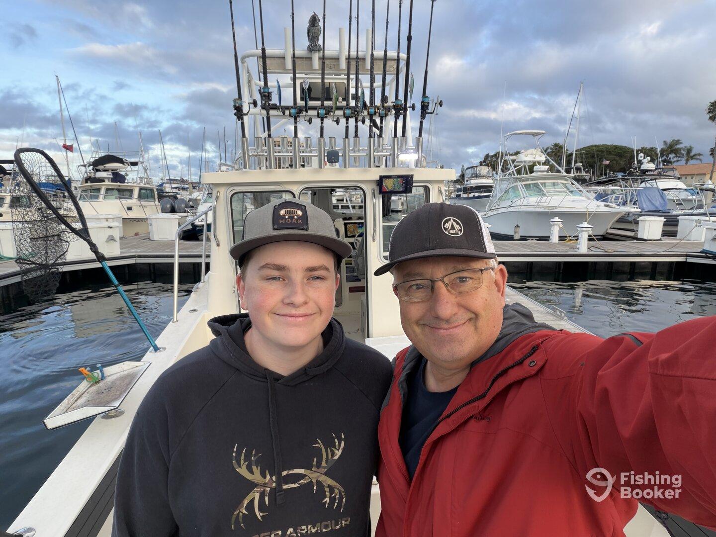 A father and son pose for a selfie on the deck of a fishing boat, surrounded by a marina filled with other boats.