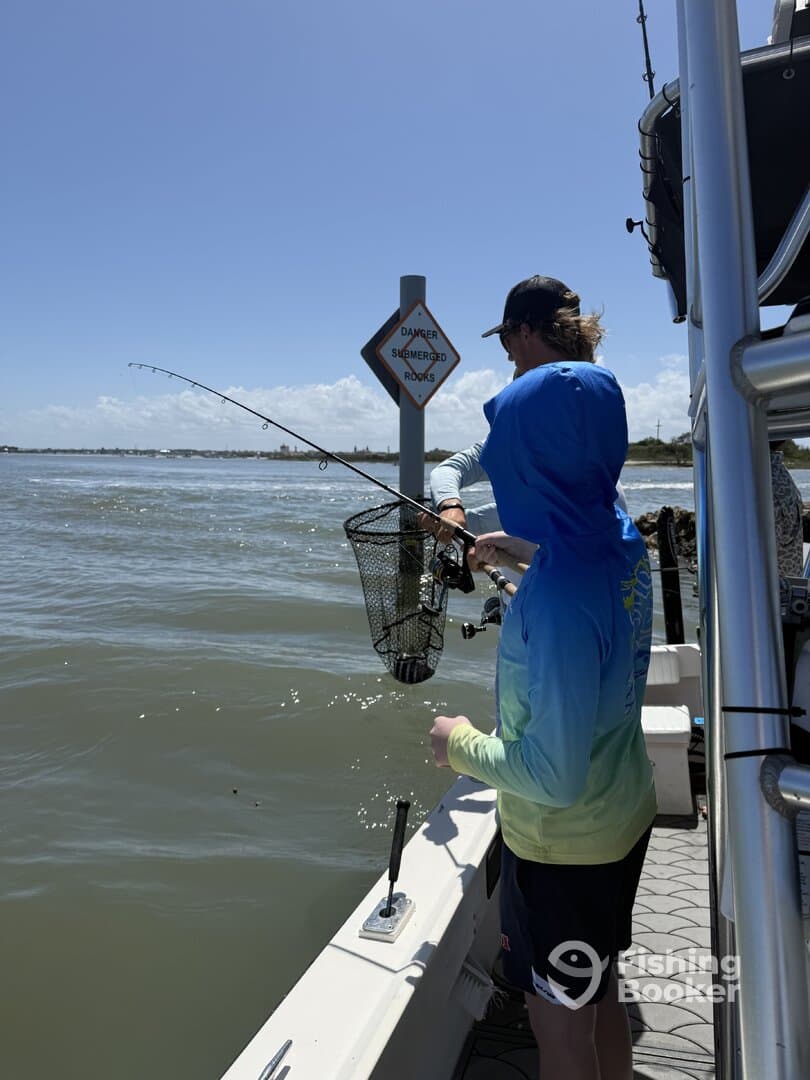 Two anglers actively fishing from a boat, with one preparing to net a catch while the other reels in. The scene captures the excitement of fishing in a coastal environment.