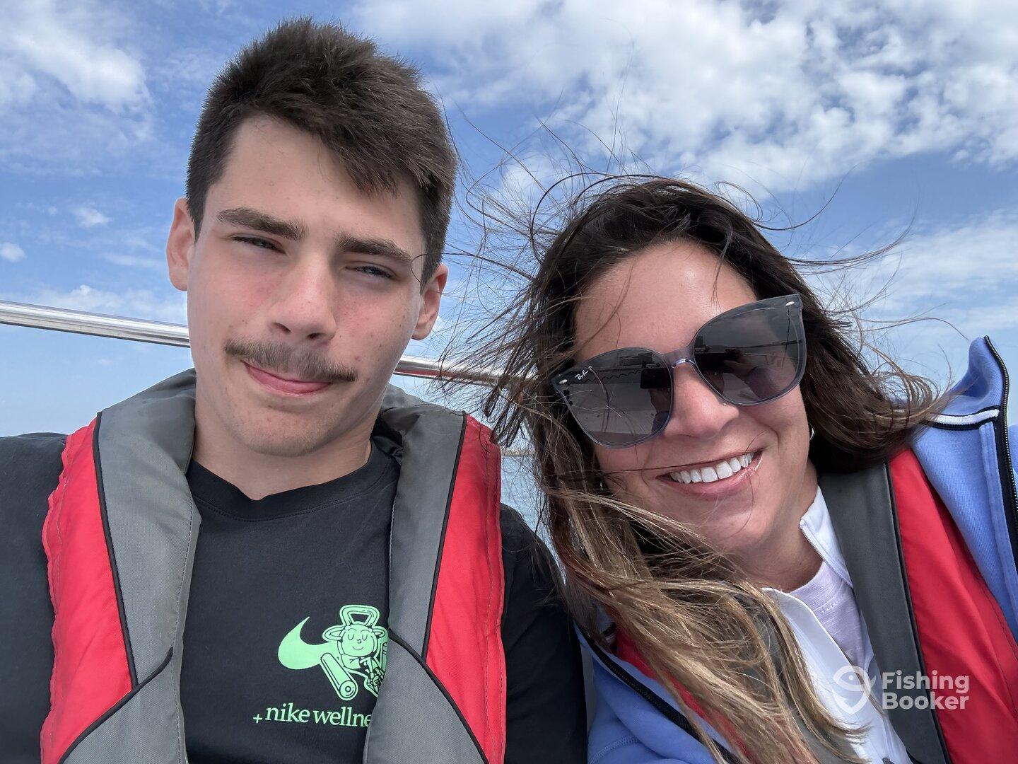A young man and an adult woman enjoying a boat ride, both wearing life jackets, with a clear sky in the background.