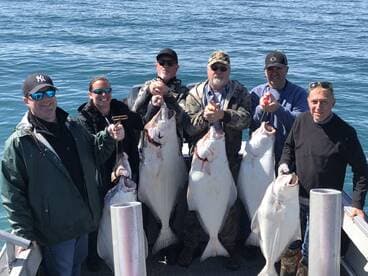 A group of six anglers proudly displaying their catch of large Halibut on a fishing charter in clear blue waters.