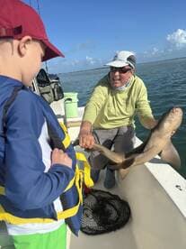 A young angler watches excitedly as an experienced fisherman holds up a caught shark aboard a fishing boat, showcasing a memorable fishing experience.