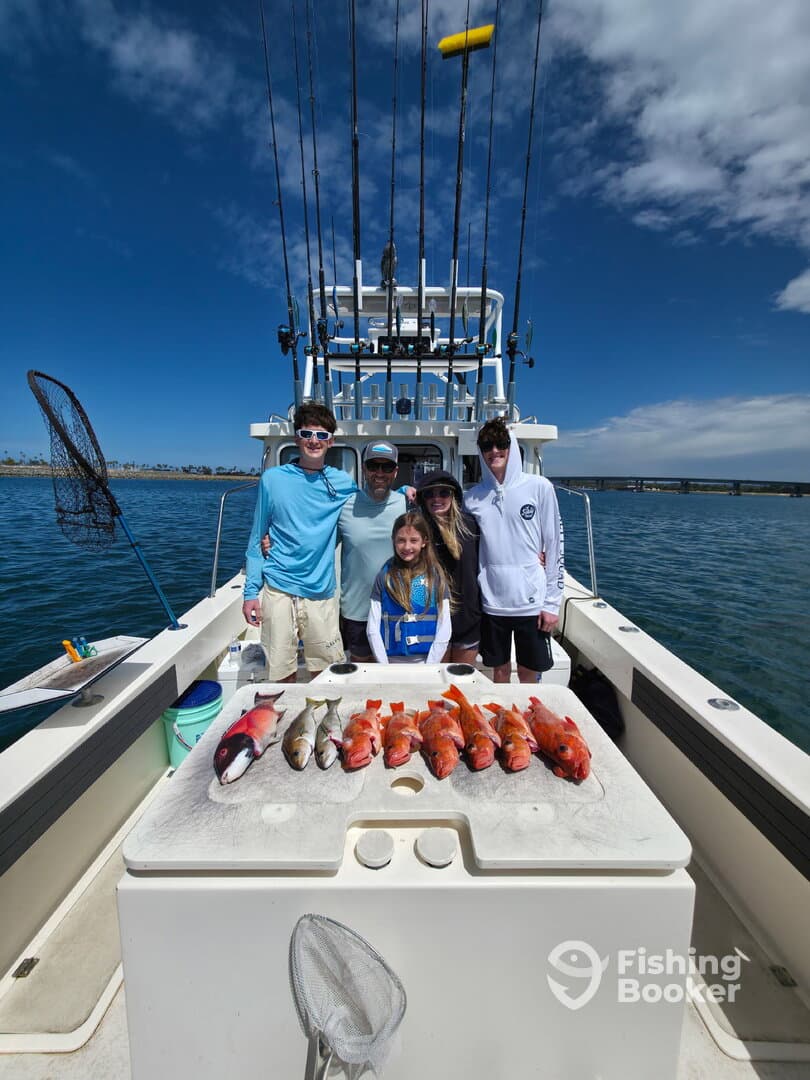 A family poses proudly on a fishing charter with a variety of fish, including Snapper and Grouper, displayed on the cleaning table against a scenic backdrop.