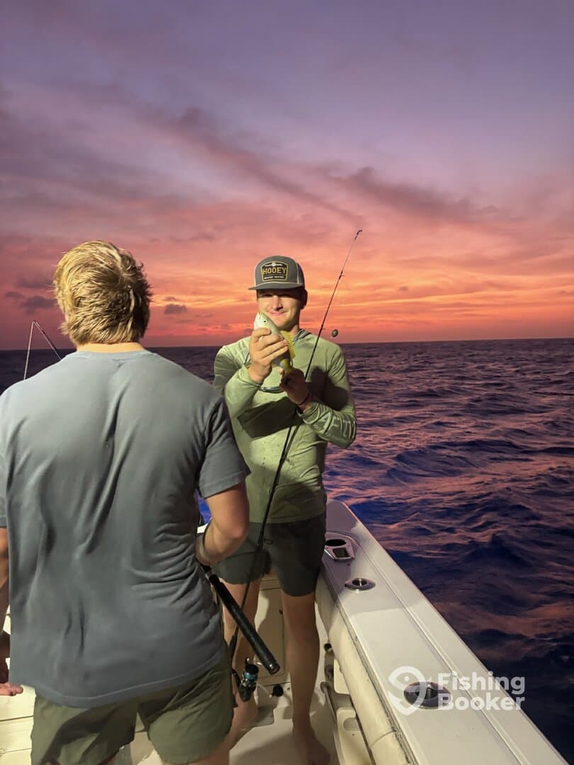 Two anglers enjoying a sunset fishing trip, with one proudly displaying a caught fish on a boat.