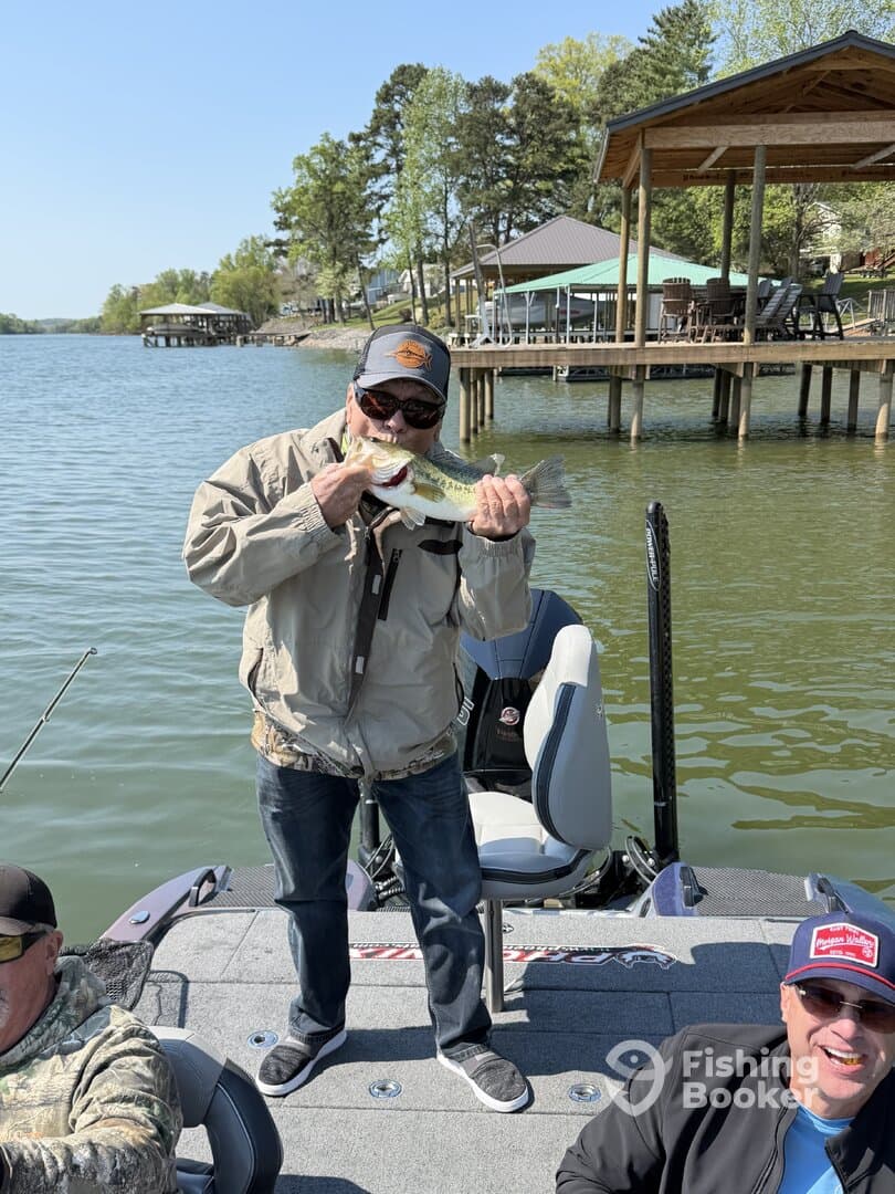 An angler proudly displaying a large Largemouth Bass while fishing from a boat on a sunny day, with a scenic lakeside backdrop.