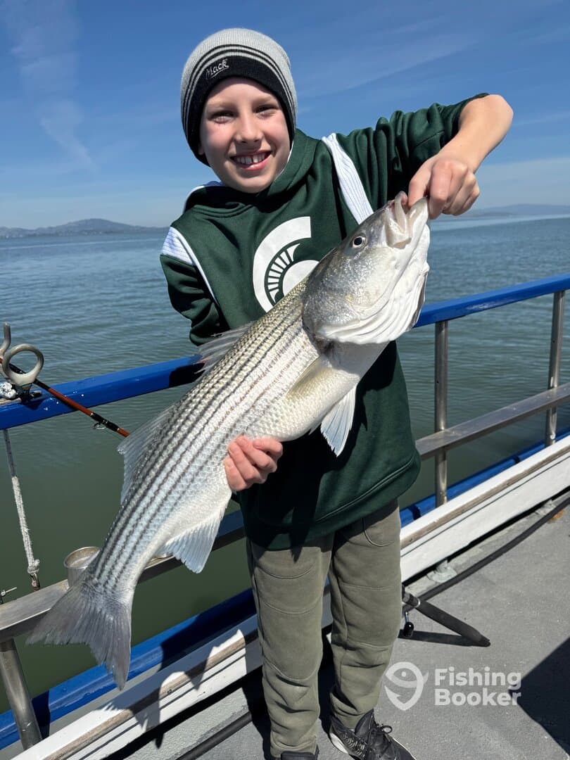 A young angler proudly displaying a large Striped Bass while fishing on a boat, with a scenic water backdrop.