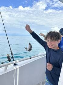 A young angler proudly displaying a freshly caught fish while fishing on a boat, with a scenic ocean backdrop.