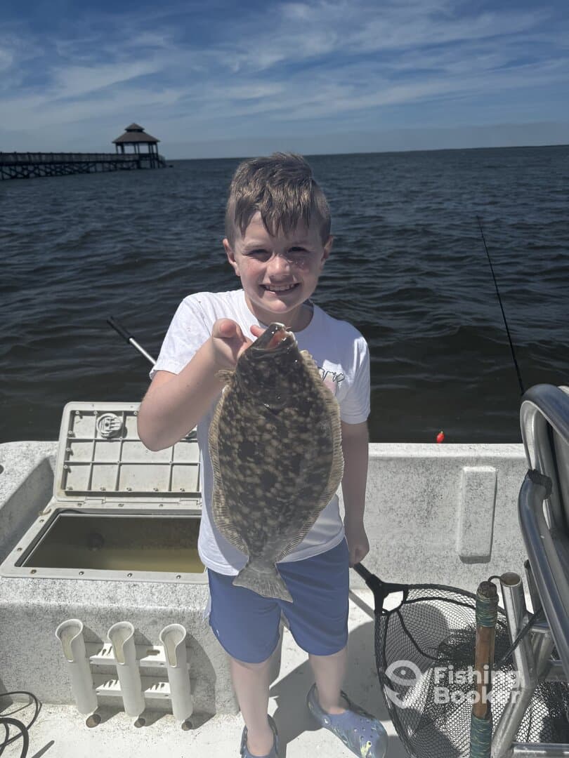 A young angler proudly displaying a Flounder caught while fishing from a boat, with a scenic pier in the background.