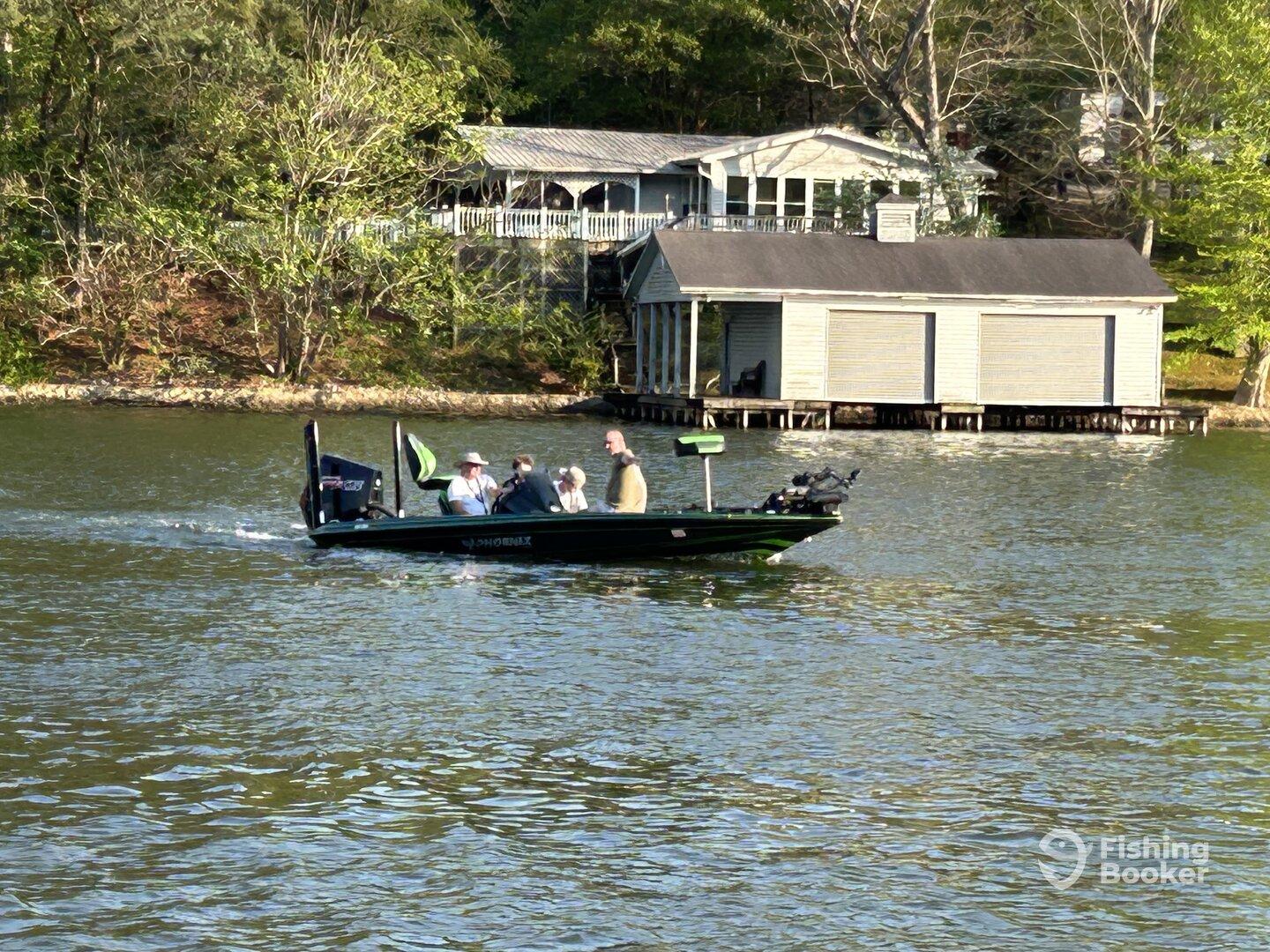 A group of anglers enjoying a day on the water in a Phoenix bass boat, cruising past a lakeside cabin.