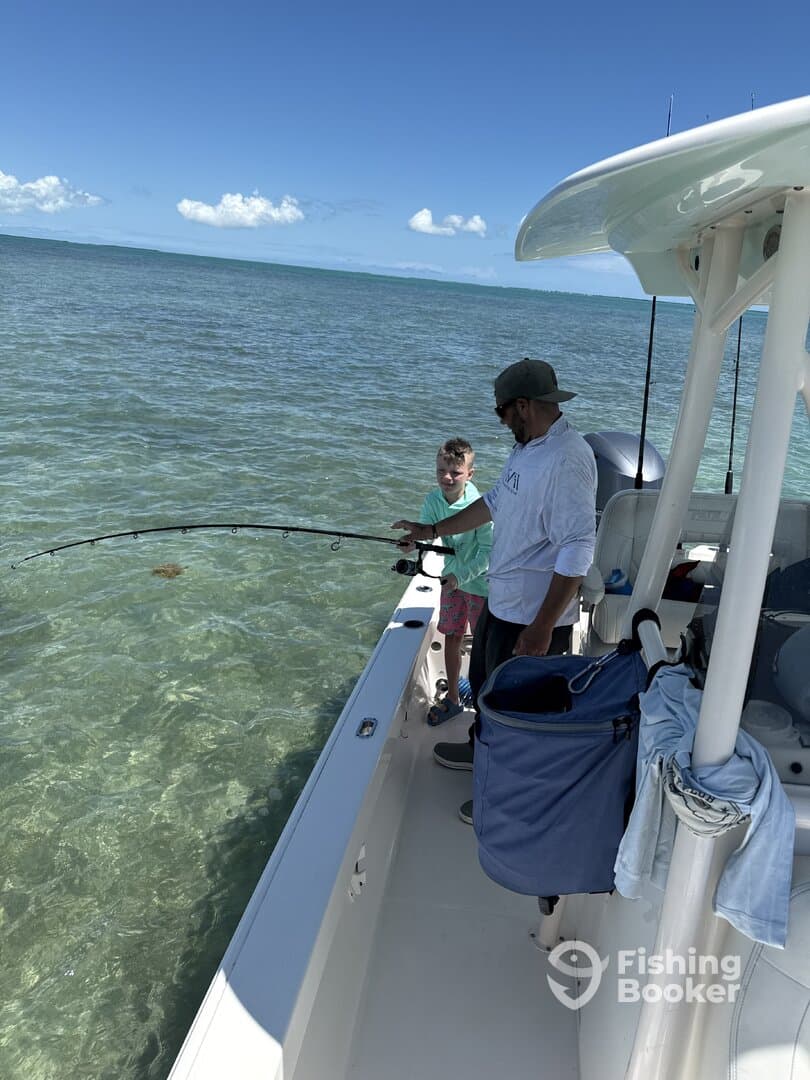 A young boy and an adult are fishing together on a boat, with clear water and a sunny sky in the background, creating a perfect fishing environment.
