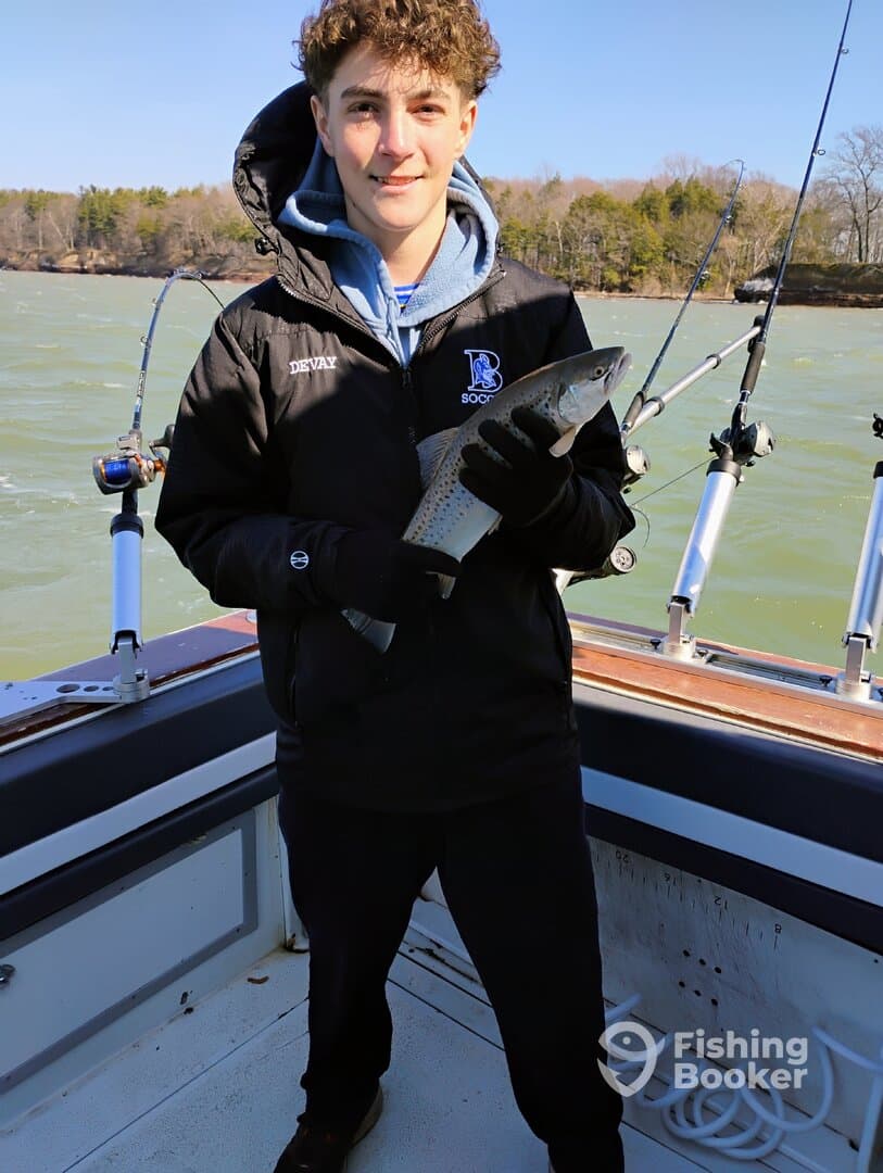 Young angler proudly holding a trout while fishing on a boat, showcasing a successful catch on a scenic day.