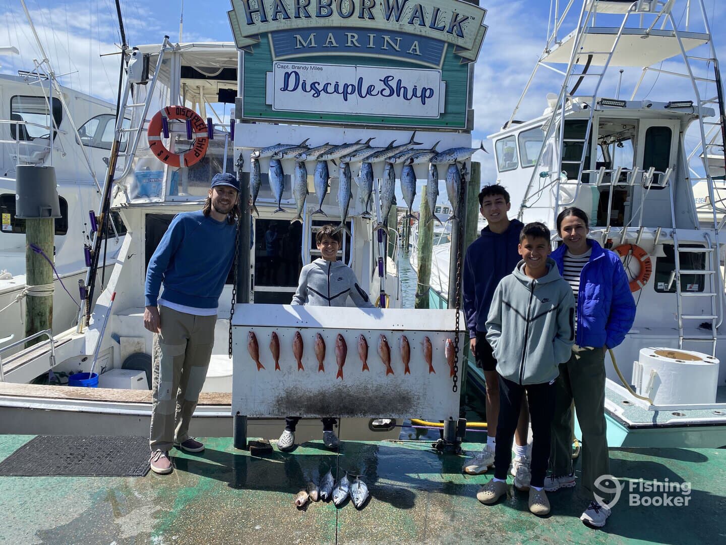 A group of young anglers proudly posing with their catch of various fish, including Snapper, displayed on a catch board at Harborwalk Marina.
