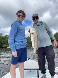 A young angler and an adult proudly display a large Redfish while fishing in a scenic coastal environment.
