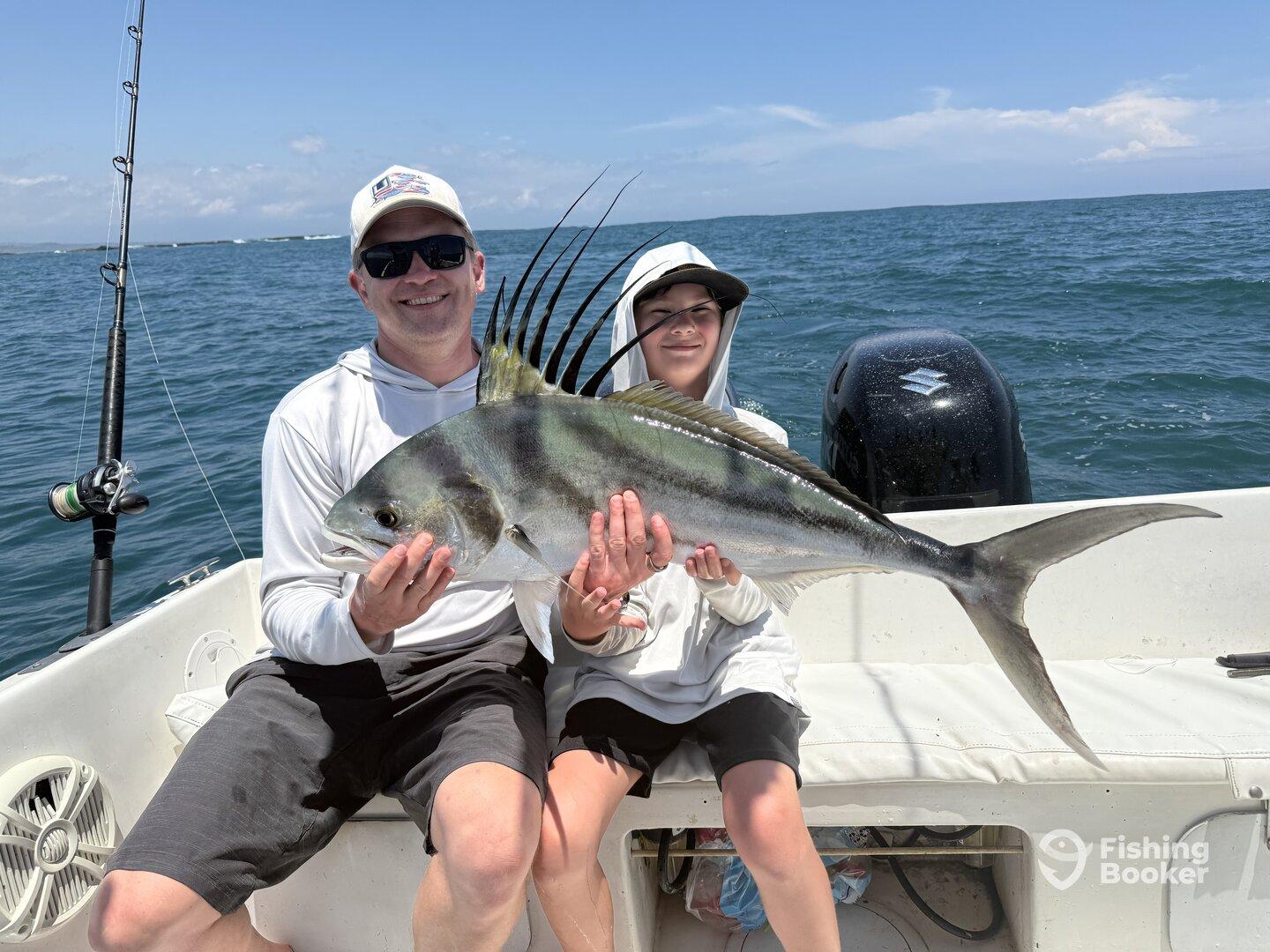 A proud father and son duo displaying a large Roosterfish while fishing on a boat, enjoying a sunny day on the water.