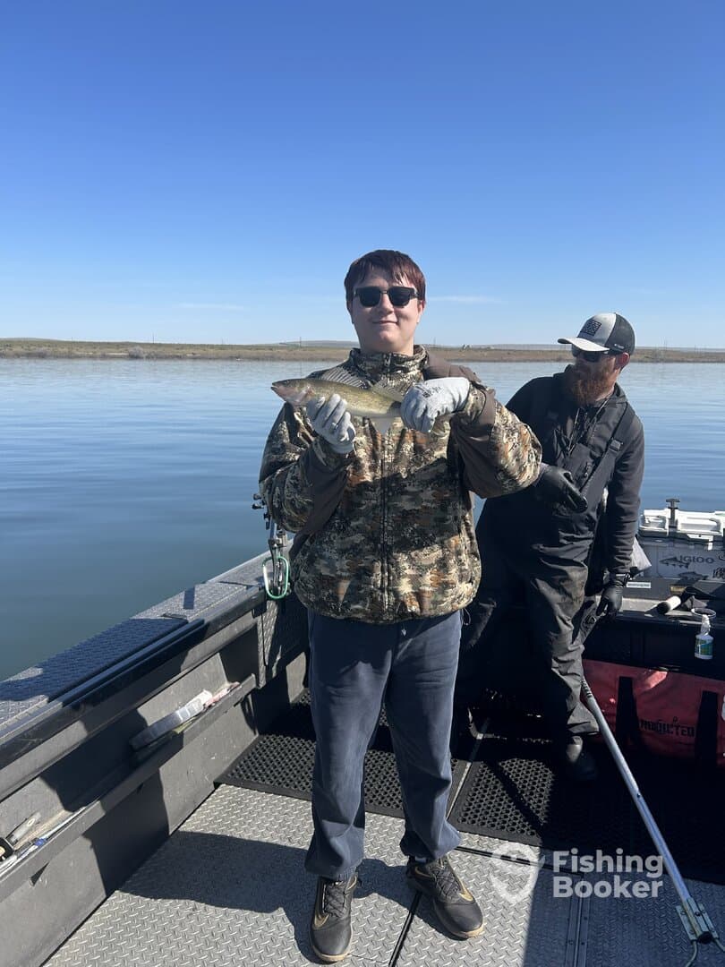 A young angler proudly displaying a caught fish while fishing on a boat, with a clear blue sky and calm waters in the background.