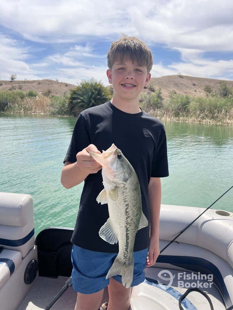 A young angler proudly displaying a large Largemouth Bass while fishing from a boat in a scenic lake environment.