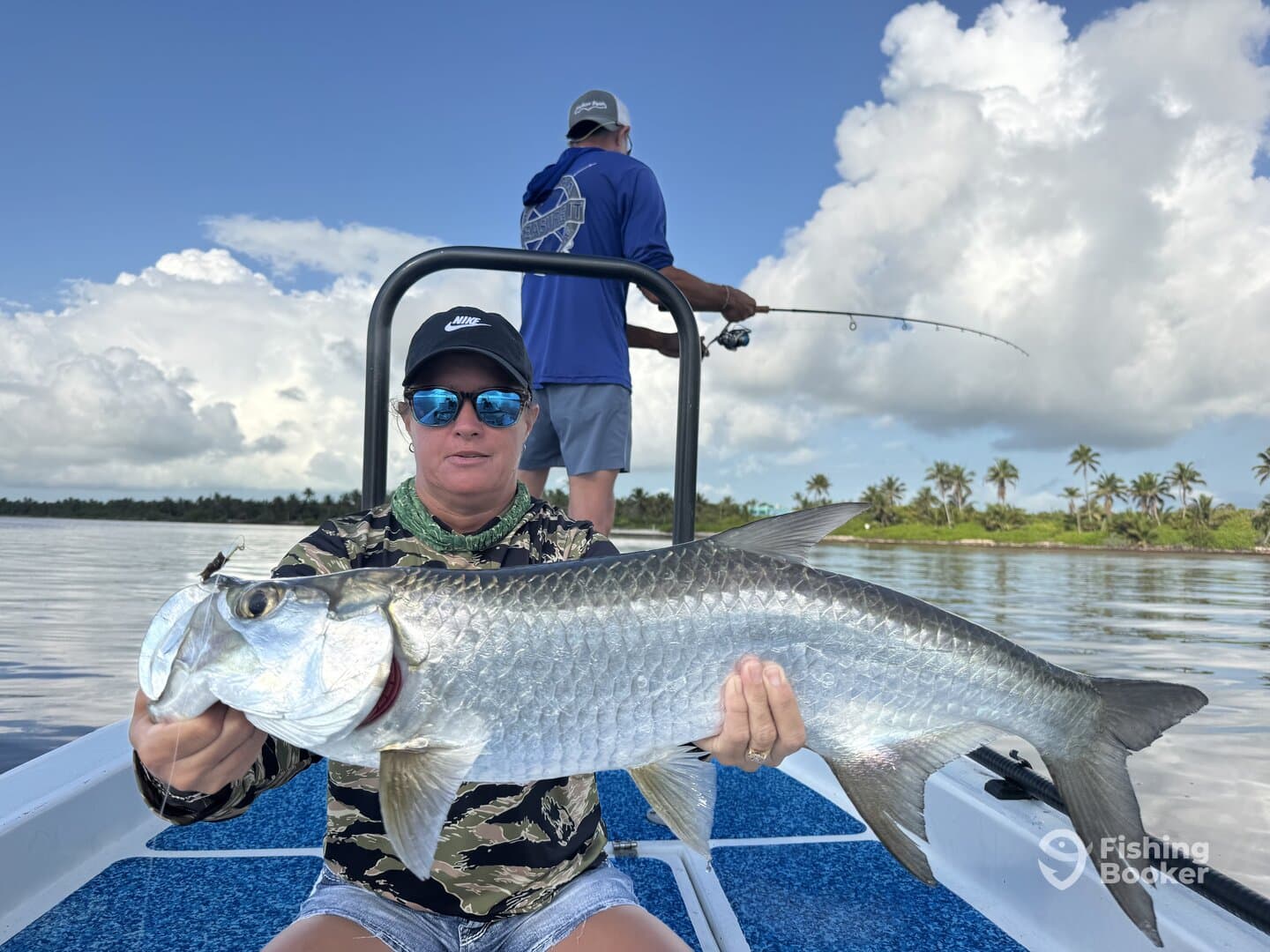 A woman proudly displays a large Tarpon while fishing on a boat in a tropical setting, with another angler in the background.