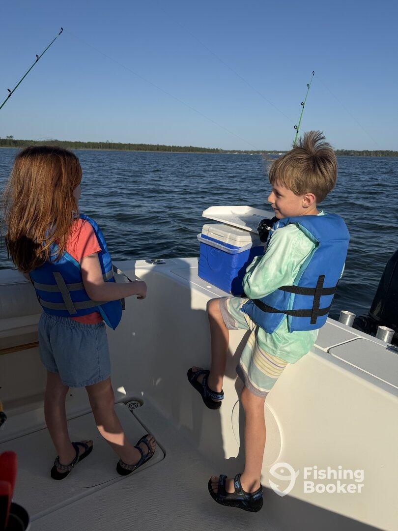 Two young anglers in life jackets enjoying a fishing trip on a boat, with rods in hand and a cooler nearby.