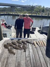 Two anglers proudly posing next to a catch of flounder and redfish displayed on a dock after a successful fishing trip.