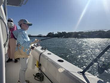A young angler casting a line from a boat in a scenic fishing environment, with a pier visible in the background.