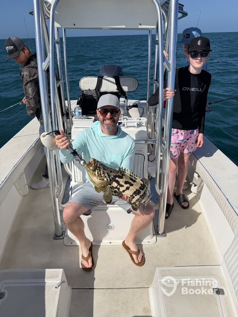 A proud angler holding a large Goliath Grouper on a fishing boat, surrounded by family members enjoying a day on the water.
