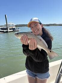 A smiling angler proudly displaying a Redfish while on a fishing trip, with a scenic backdrop of the water and a boat.