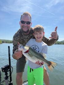 A young boy and his father proudly display a large Largemouth Bass while fishing on a sunny day.