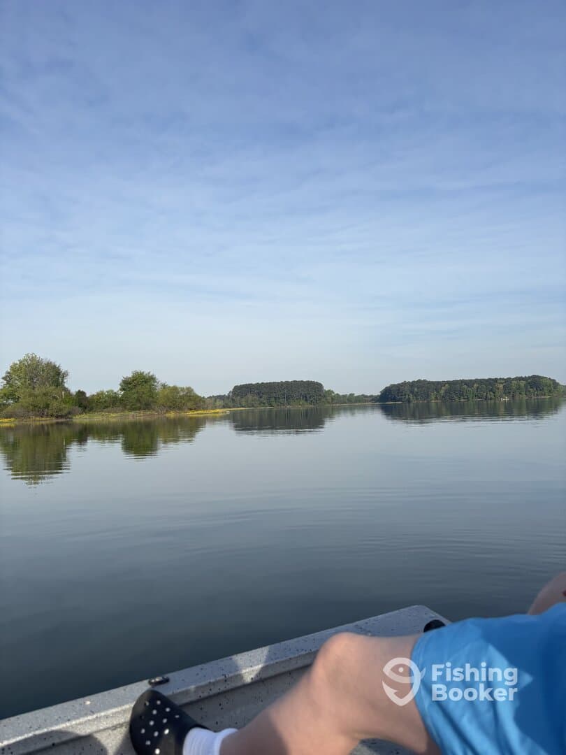 A serene view from inside a small boat, showcasing calm waters and lush greenery, with a person's leg visible in the foreground.