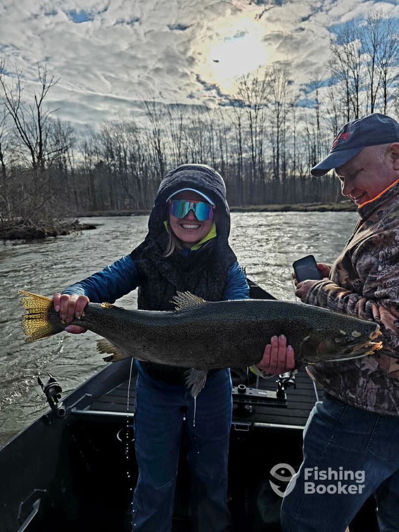 A smiling angler proudly displays a large trout while fishing on a river, showcasing a fun day out with friends.