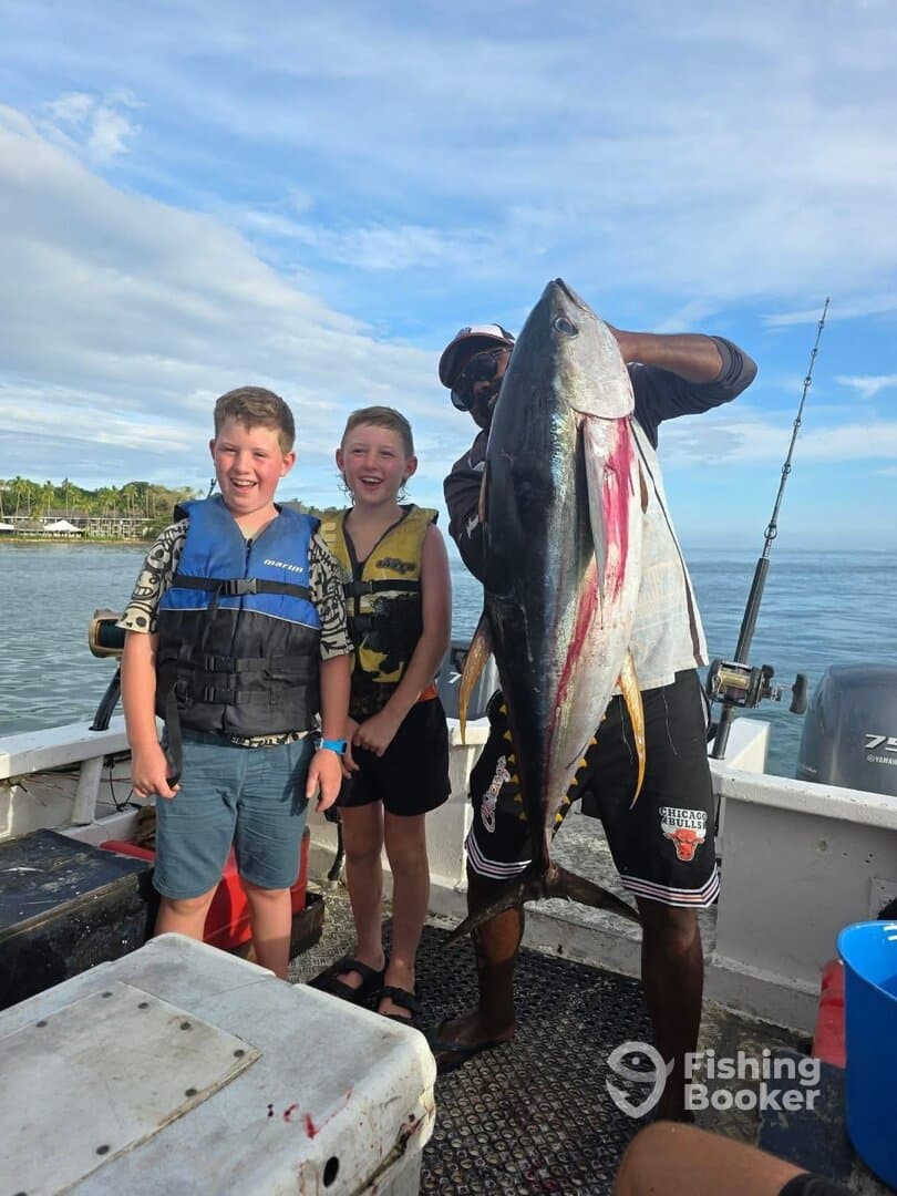 Two young anglers proudly posing with a large Tuna on a fishing charter, showcasing their successful catch in a scenic coastal environment.