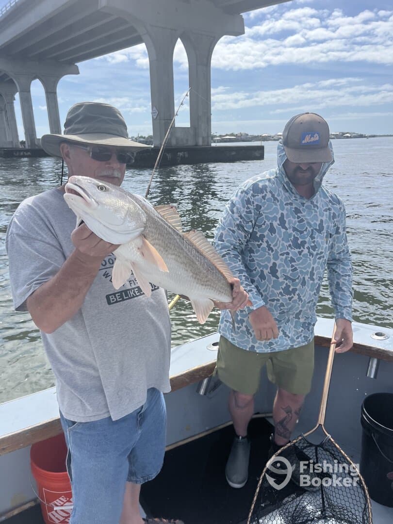 Two anglers proudly displaying a large Redfish while fishing under a bridge, showcasing a successful day on the water.