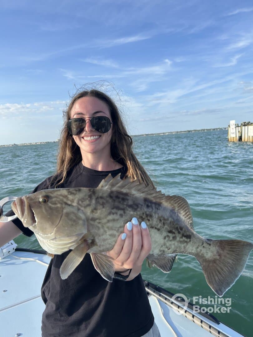 A young angler proudly displaying a large Grouper while fishing on a boat, with a scenic backdrop of the water and sky.