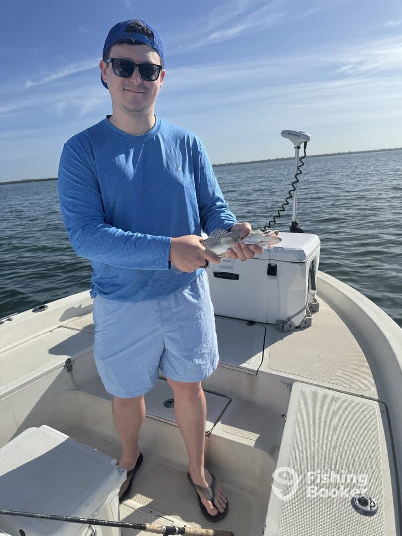 Angler holding a Speckled Trout on a boat, enjoying a sunny day on the water.