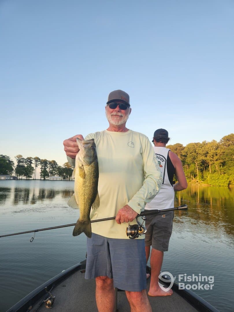 An angler proudly displaying a large Largemouth Bass while fishing on a serene lake, accompanied by another fisherman in the background.