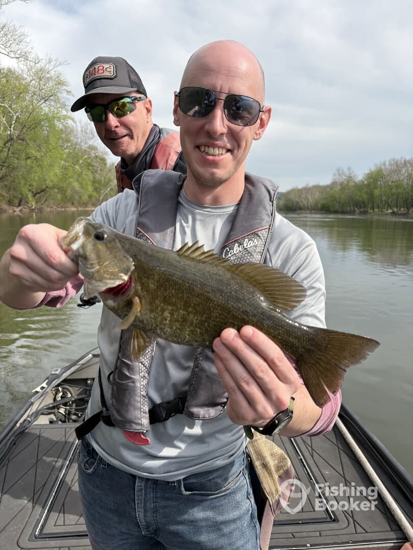 Angler proudly displaying a large Largemouth Bass while fishing on a river, with a fellow fisherman in the background.