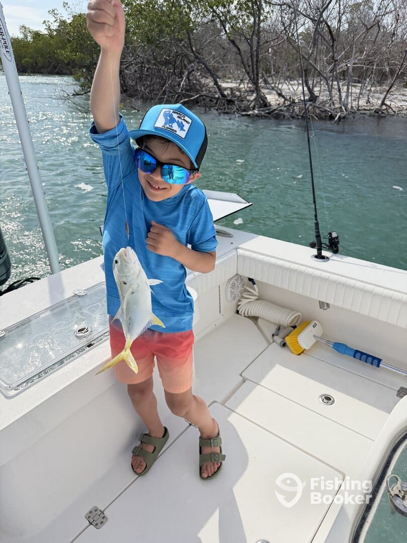 A young angler proudly displaying a small fish while fishing from a boat in a mangrove area.