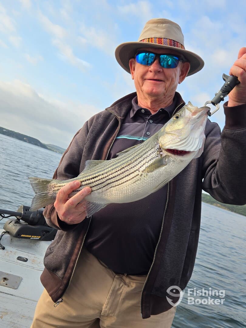 An angler proudly displaying a striped bass while fishing on a boat, showcasing a successful catch in a scenic waterway.