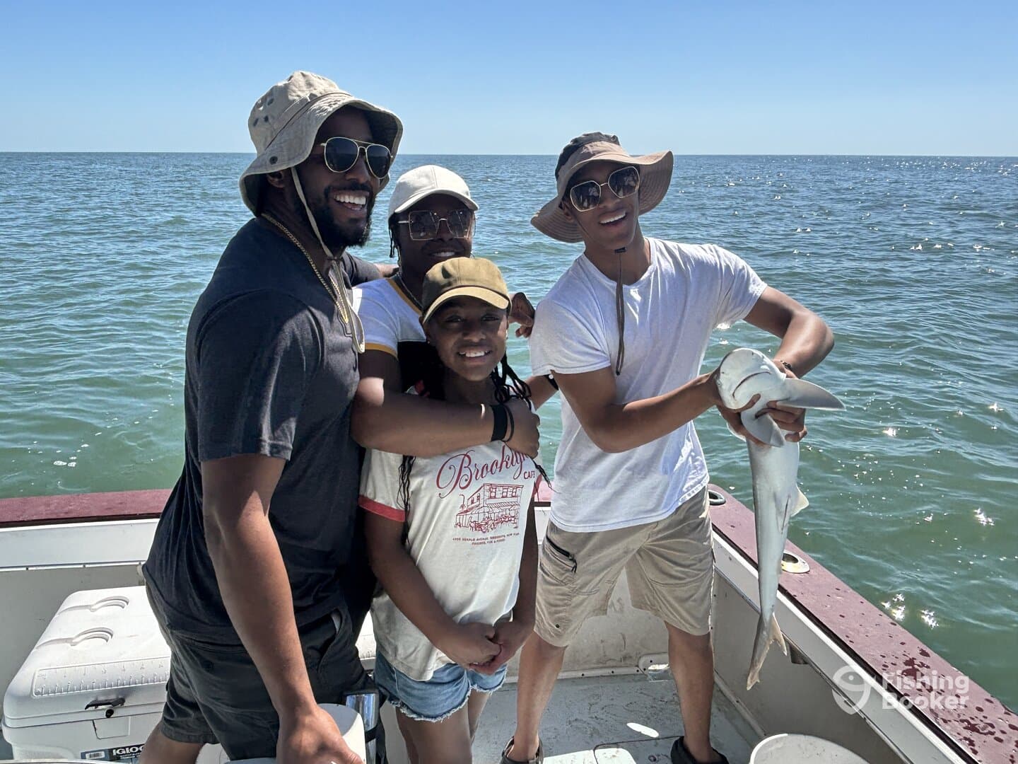 A group of four anglers enjoying a successful fishing trip, with one proudly displaying a caught shark on a boat in sunny waters.